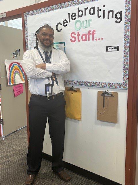  Mr. Miller crosses his arms and smiles to camera from inside a school building.